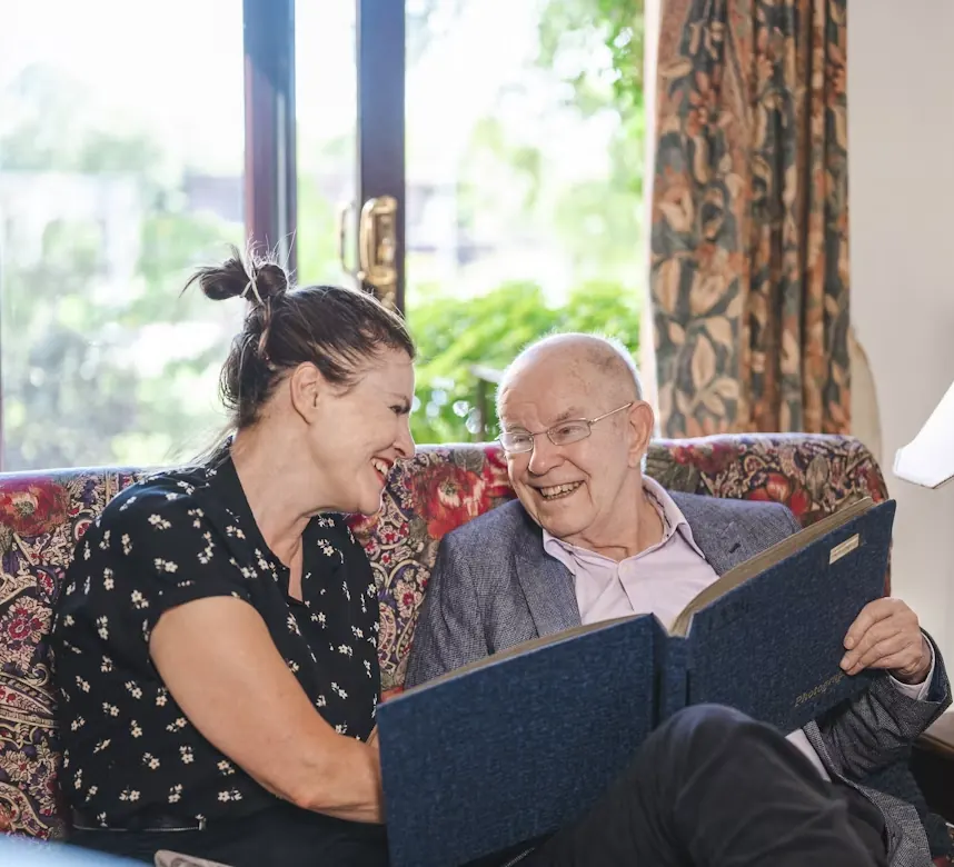 An older man and a woman laughing together as they look through a folder of papers on the couch