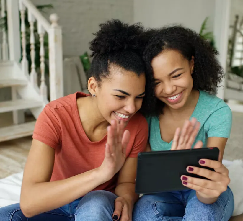 Two young women smiling and waving at someone on a tablet video call